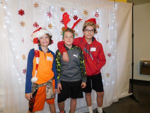 3 kids posing in front of a photo backdrop with white lights and gold and red snowflakes