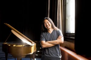 Picture of a man with long hair standing in front of a piano with his arms crossed.